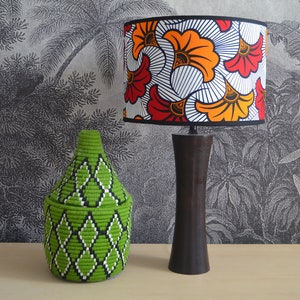 May include: A table lamp with a dark brown wooden base and a cylindrical lampshade featuring a vibrant orange, red, and white floral pattern, positioned next to a lime green woven basket with a lid and a black and white diamond geometric design.