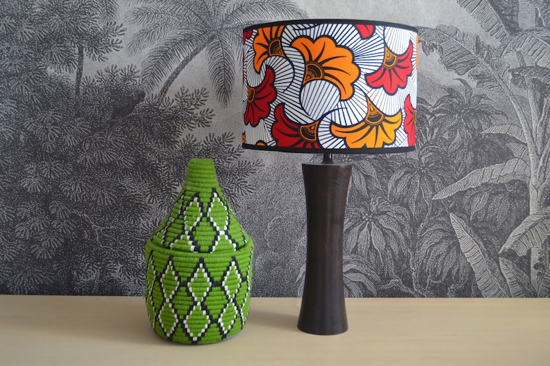 May include: A table lamp with a dark brown wooden base and a cylindrical lampshade featuring a vibrant orange, red, and white floral pattern, positioned next to a lime green woven basket with a lid and a black and white diamond geometric design.