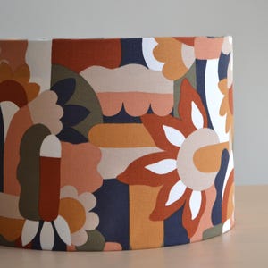 K&ouml;nnte beinhalten: Ein zylindrischer Lampenschirm mit einem farbenfrohen geometrischen Muster. Das Muster zeigt abstrakte Formen in Orange-, Braun-, Beige-, Wei&szlig;- und Gr&uuml;nt&ouml;nen auf einem dunkelblauen Hintergrund.