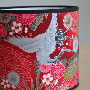 May include: Cylindrical lampshade with a vibrant red background, adorned with a detailed pattern of flowers, leaves, and a graceful crane in flight. The design is accented with black trim at the top and bottom.