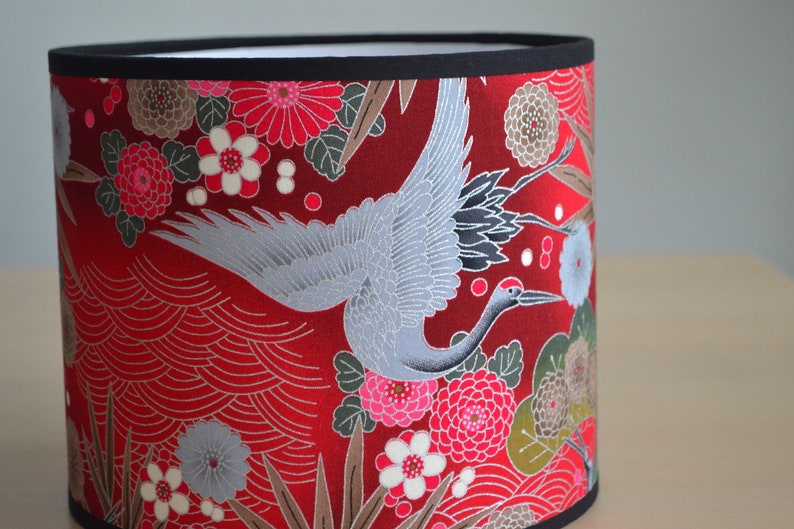 May include: Cylindrical lampshade with a vibrant red background, adorned with a detailed pattern of flowers, leaves, and a graceful crane in flight. The design is accented with black trim at the top and bottom.