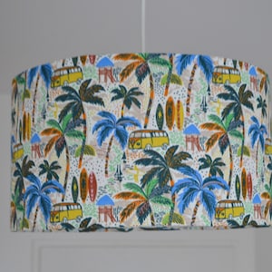Puede incluir: Una pantalla de lámpara cilíndrica blanca con un estampado tropical colorido que presenta palmeras, tablas de surf y furgonetas vintage amarillas.