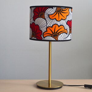 May include: A table lamp with a cylindrical lampshade featuring a bold floral pattern in red, orange, black, and white. The lamp has a slender gold-toned metal stem and a circular gold-toned base, connected to a black power cord with a switch.