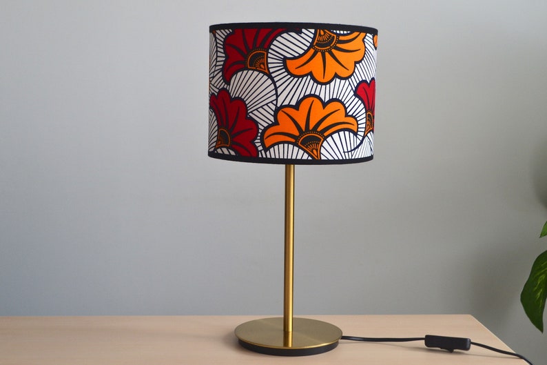 May include: A table lamp with a cylindrical lampshade featuring a bold floral pattern in red, orange, black, and white. The lamp has a slender gold-toned metal stem and a circular gold-toned base, connected to a black power cord with a switch.