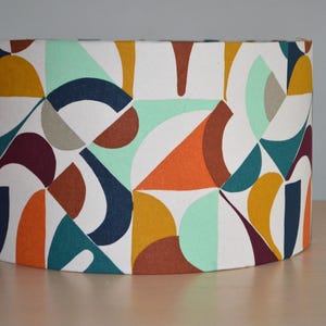 Peut inclure: Abat-jour cylindrique avec un motif géométrique. Le design présente des formes orange, sarcelle, marine, jaune moutarde et bordeaux sur fond blanc. L'abat-jour est en tissu.