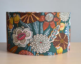 May include: A cylindrical lampshade featuring a dark green background with a dense, colorful botanical pattern. The design includes stylized flowers and leaves in shades of burnt orange, mustard yellow, white, and light blue with intricate line work.