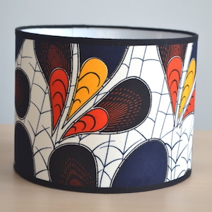 May include: A cylindrical lampshade with a black trim and a white background. The lampshade features a colourful geometric pattern with red, yellow, and black accents.