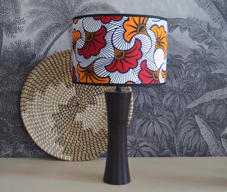 May include: A table lamp with a dark brown, smooth, cylindrical base that tapers slightly toward the center. The drum-shaped lampshade features a vibrant, repeating pattern of stylized orange and red floral shapes on a white background.
