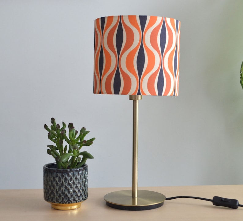 May include: A gold table lamp with a drum shade featuring a retro orange and blue wavy pattern. The lamp is on a wooden table with a small potted succulent plant.