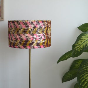 May include: A floor lamp with a gold metal stand and a drum lampshade with a pink, brown, and gold geometric pattern.