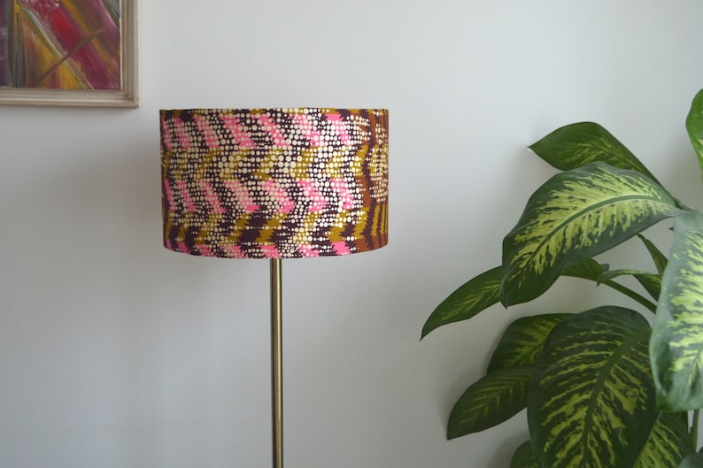 May include: A floor lamp with a gold metal stand and a drum lampshade with a pink, brown, and gold geometric pattern.