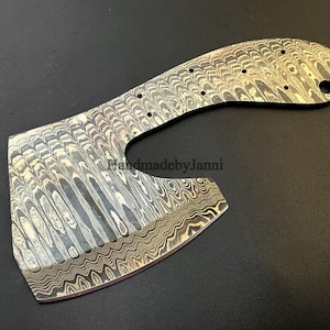 Puede incluir: Un primer plano de una cabeza de hacha de acero Damasco hecha a mano con un patrón único. La cabeza del hacha tiene una hoja curva y un mango redondeado. El texto "HandmadebyJanni" es visible en la cabeza del hacha.