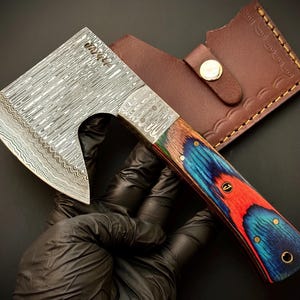 May include: A small, decorative axe with a patterned steel blade and a colorful wooden handle. The handle features red, blue, and green hues with brass accents. A brown leather sheath with a snap closure is included.