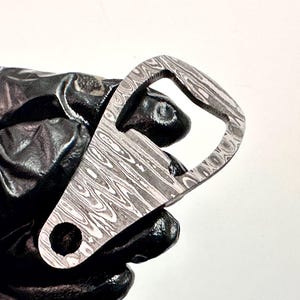 Könnte beinhalten: Ein silberner Flaschenöffner mit einem gemusterten Design. Der Öffner ist aus Metall und hat eine glatte, polierte Oberfläche.