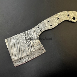 Puede incluir: Una cabeza de hacha en blanco hecha de acero Damasco con un acabado estampado. La cabeza del hacha tiene un mango con un agujero para un cordón. El texto "Handmade by Janni" es visible en la cabeza del hacha.