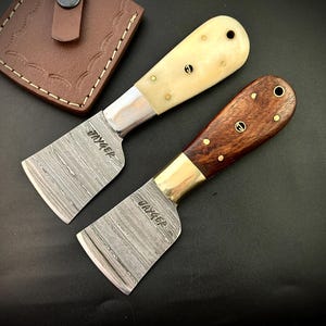 May include: Two small leather working knives with Damascus steel blades and different handle materials. The knife on the left has a white handle and the knife on the right has a dark wood handle. Both knives have the word "VAYGER" engraved on the blade.