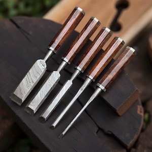 Puede incluir: Juego de cinco formones de madera con hojas de acero estampadas y mangos de madera oscura. Los mangos tienen detalles metálicos plateados y están grabados con el texto "JAYGER". Los formones están dispuestos sobre una superficie de madera oscura.