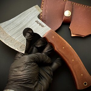 May include: A small, hand-forged axe with a Damascus steel blade and a brown wooden handle. The axe has a leather sheath with a decorative stitching pattern. The blade is engraved with the word "JAEGER".
