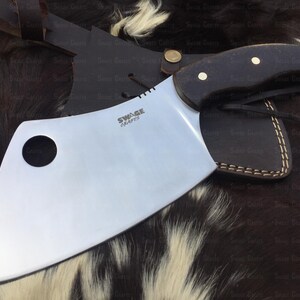 May include: A large, curved, stainless steel cleaver with a black wooden handle. The blade has a hole in the center and the text "SWAGE CRAFTS" is engraved on the blade. The cleaver is resting on a brown and white fur surface.