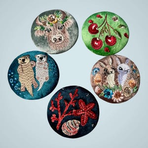 May include: Five circular embroidered patches featuring distinct designs: a cow with a floral crown, a branch of red cherries, two sea otters, two rabbits surrounded by flowers, and a red coral branch with a starfish and a seashell.