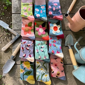 May include: A selection of floral-patterned socks in shades of pink, blue, green, and yellow, presented on a wooden surface. The socks showcase diverse flower designs and are displayed with gardening tools and a watering can.