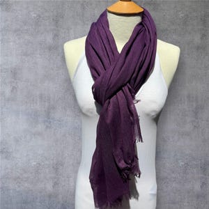 May include: A dark purple scarf displayed on a mannequin. The scarf is made of a lightweight material with a textured finish and fringed edges. The scarf is knotted loosely around the neck.