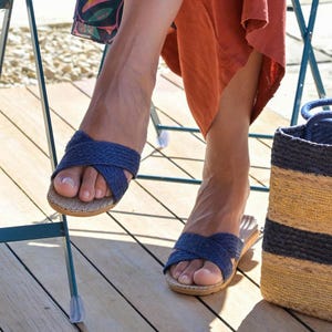 May include: A pair of blue woven slide sandals with a criss-cross strap design. The sandals are worn by a person sitting on a wooden deck.