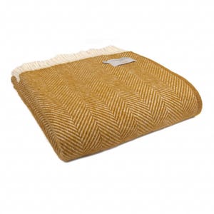 May include: Mustard yellow herringbone pattern throw blanket with a cream-colored fringe. The blanket is folded, showcasing the textured weave and a small tag. A cozy home decor item.