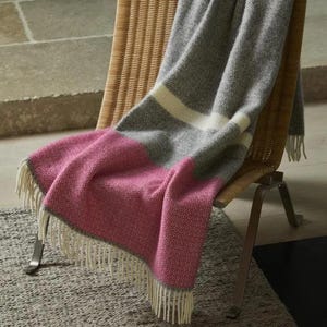 May include: A pink, grey and white striped throw blanket with a fringed edge, draped over a wicker chair. The blanket is made of a woven material. The chair has metal legs.