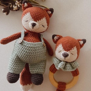 May include: Two crocheted fox toys. The larger fox is wearing green overalls and has a brown tail. The smaller fox is attached to a wooden ring and has a brown tail. Both foxes have closed eyes and brown noses.