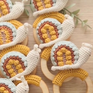 May include: Crocheted snail baby toys with wooden rings. The snails have cream-colored bodies, yellow and blue shells, and are arranged on a wooden surface. The toys are handmade and suitable for babies.