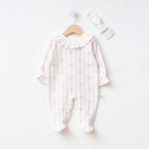 May include: A white baby romper with a pink bow pattern and a ruffled collar hangs on a wooden hanger. A matching headband with a bow is also visible. The romper has long sleeves and footed bottoms.