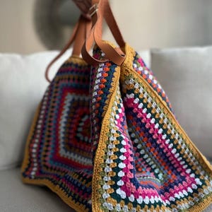 May include: A colorful, handmade crochet tote bag with a square pattern. The bag features a variety of colors including pink, blue, orange, and white. It has brown leather straps and a mustard yellow border.