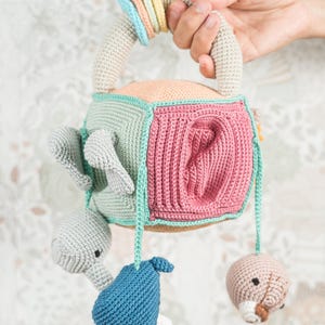 May include: A crocheted baby toy with a cube shape and a handle. The cube has different colored panels and features a variety of crocheted animal figures hanging from it. The toy is made in pastel colors, including pink, blue, and green.