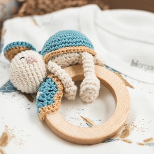 May include: A crocheted blue and tan turtle toy attached to a wooden teething ring. The toy is lying on a white baby onesie with a brown and gold print.