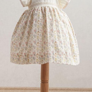 May include: A white baby dress with a floral pattern, displayed on a wooden mannequin. The dress features short, ruffled sleeves and a delicate lace trim. The skirt is adorned with a colorful floral print.