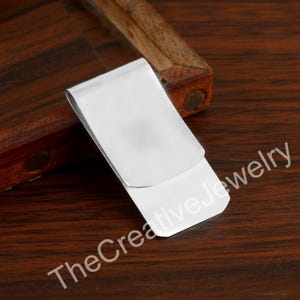 May include: A shiny, silver-colored money clip. The clip is rectangular with a curved top and is resting on a dark wooden surface. The text "TheCreativeJewelry" is printed on the bottom right.