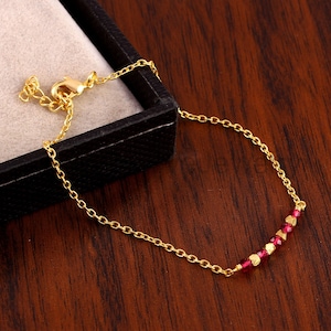 May include: A delicate gold bracelet with a chain design. The bracelet features small, faceted red beads and gold beads. The bracelet is displayed on a dark wooden surface.