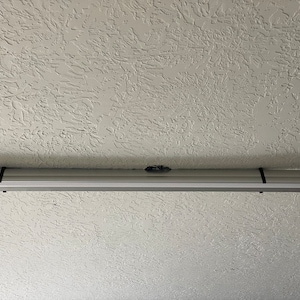 May include: A white ceiling-mounted projector screen with a black pull cord. The screen is retracted and the ceiling has a textured finish.