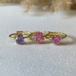 Könnte beinhalten: Drei goldene Ringe mit bunten Edelsteinen. Die Ringe haben einen lila, rosa und rosa herzförmigen Stein.