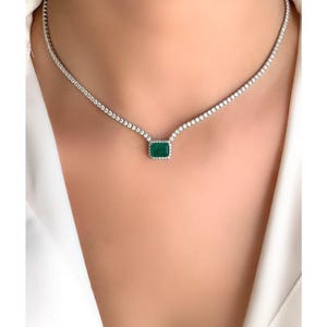 Puede incluir: Un collar de plata con un solo colgante de piedra preciosa verde esmeralda rodeado de piedras preciosas más pequeñas y transparentes. El collar es una cadena delicada con piedras preciosas transparentes.