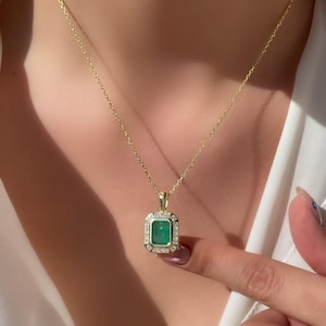 May include: A gold chain necklace with a square emerald green pendant set in a gold frame with small clear stones around the edge.