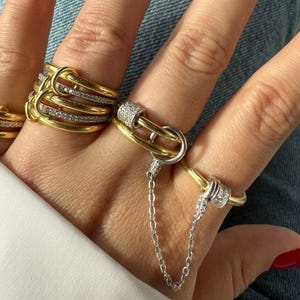 Könnte beinhalten: Eine Sammlung von Gold- und Silberringen mit verschiedenen Designs. Einige Ringe haben ein Kettenelement, während andere mit kleinen, funkelnden Steinen verziert sind. Die Ringe werden auf einer Hand mit rotem Nagellack präsentiert.