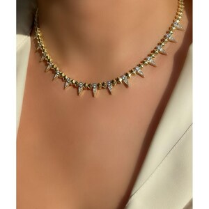 Puede incluir: Un collar gargantilla de oro con pequeñas piedras transparentes y puntas puntiagudas. El collar está puesto en el cuello de una modelo.