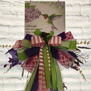 May include: A purple, green, and pink bow with a white lace ribbon hangs from a sign that reads "although we are apart, you are in my heart". A hummingbird with a long beak is perched on a purple flower.