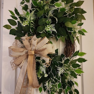 May include: A green and white leafy wreath with a brown burlap and white striped bow. The wreath is hanging on a white door.