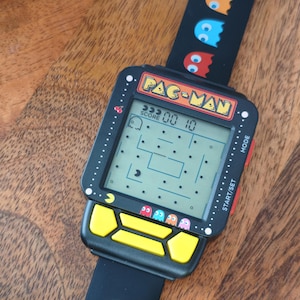 Könnte beinhalten: Eine schwarze Digitalarmbanduhr mit einem Pac-Man-Spielbildschirm und dem Schriftzug PAC-MAN oben. Die Uhr hat ein quadratisches Gehäuse, gelbe Steuertasten und ein schwarzes Silikonarmband, das mit bunten Geisterfiguren aus dem Spiel verziert ist.