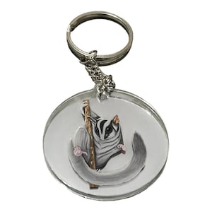 May include: A circular clear acrylic keychain featuring a detailed illustration of a grey and white sugar glider clinging to a brown vertical branch. The animal has large dark eyes, pointed ears, and a long tail. It is attached to a silver metal ring.