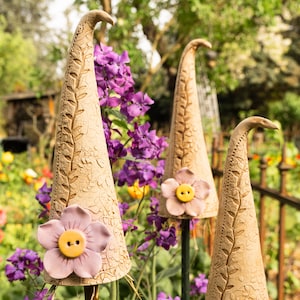 Könnte beinhalten: Drei Keramik-Gartenpfähle in Form von hohen, spitzen Hüten mit Blumendesign. Jeder Pfahl hat eine rosa Blume mit einem gelben Knopf in der Mitte. Die Pfähle stehen in einem Garten mit lila Blumen im Hintergrund.
