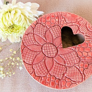 Könnte beinhalten: Eine runde, korallenfarbene Keramikschale mit herzförmigem Ausschnitt. Die Schale hat ein erhabenes Blumendesign mit komplizierten Mustern. Eine weiße und gelbe Blume und kleine weiße Blumen sind im Hintergrund.